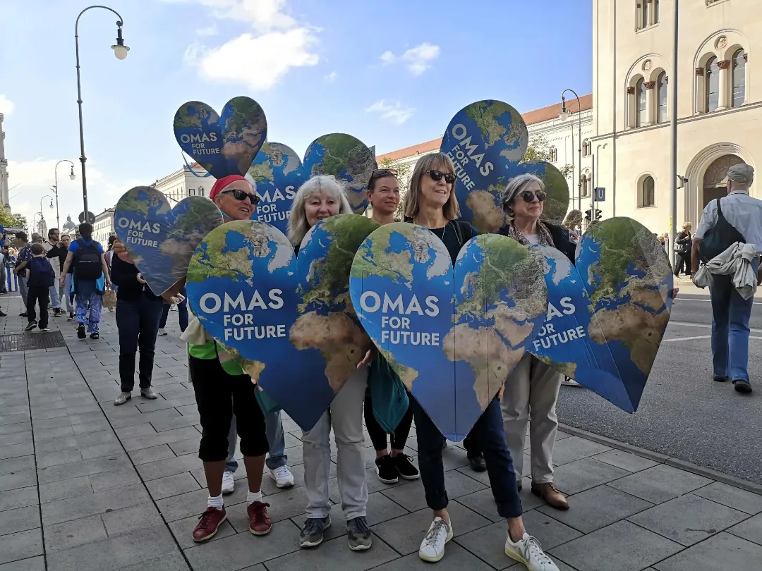 Eine Gruppe älterer Frauen steht auf einer Straße in der Stadt und hält große herzförmige Schilder mit einer Weltkarte und der Aufschrift OMAS FOR FUTURE in die Höhe, um an einem Klimaprotest teilzunehmen.