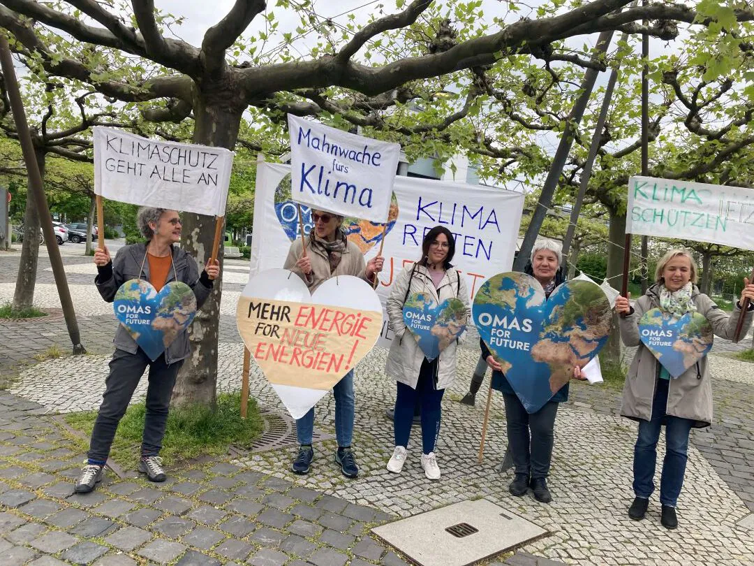 Fünf Menschen stehen unter freiem Himmel mit Klimaprotestschildern in deutscher Sprache, herzförmigen Omas for Future-Tafeln und Bannern für den Klimaschutz unter blattlosen Bäumen auf einem Kopfsteinpflasterweg.