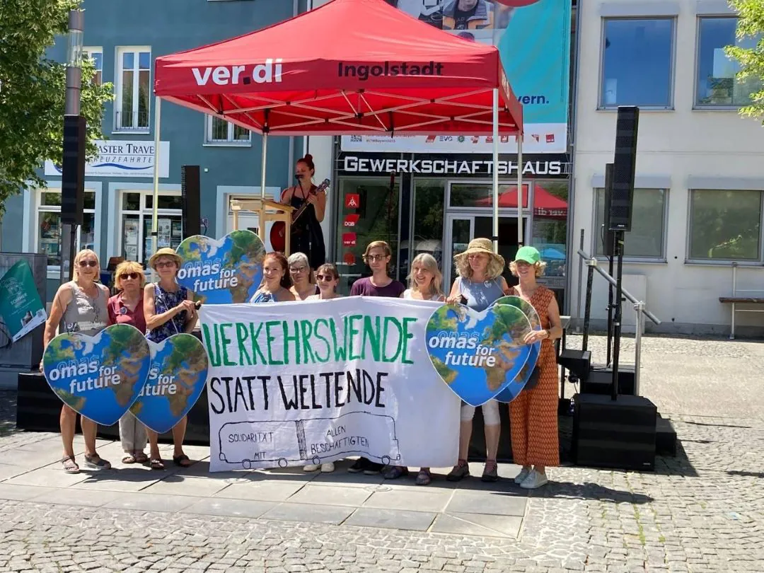 Eine Gruppe von Menschen hält ein Transparent mit der Aufschrift VERKEHRSWENDE STATT WELTENDE und omas for future unter einem roten Zelt bei einer Freiluftveranstaltung vor dem Gewerkschaftshaus in Ingolstadt. Eine Frau spricht an einem Podium.