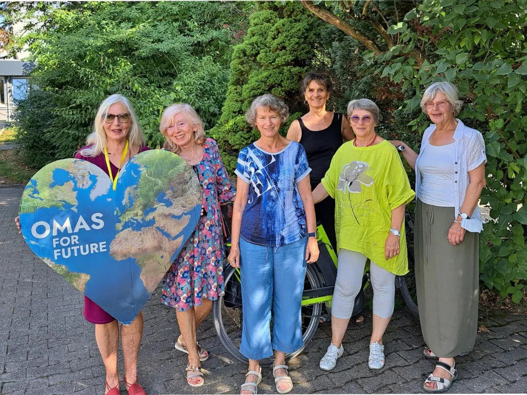 Sechs ältere Frauen stehen lächelnd im Freien; eine hält ein großes herzförmiges Schild mit einer Weltkarte und dem Text "OMAS FOR FUTURE". Im Hintergrund sind grüne Bäume und Sträucher zu sehen, und ein geparktes Fahrrad ist teilweise hinter ihnen zu erkennen.