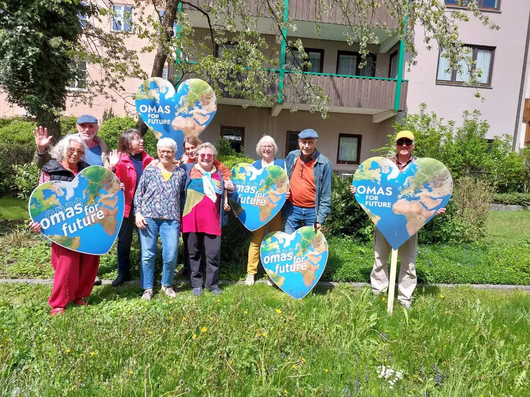 Eine Gruppe älterer Menschen steht auf einer Wiese vor einem Wohnhaus und hält große herzförmige Schilder mit der Aufschrift "Omas for Future" in Form einer Weltkugel in die Höhe, lächelt und winkt.