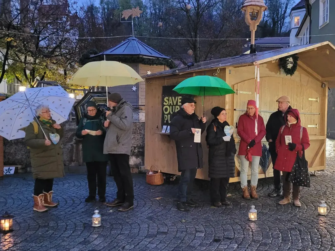 Eine Gruppe von Menschen steht bei einer Veranstaltung im Freien mit Kerzen und Regenschirmen vor einer Holzhütte. Laternen beleuchten den kopfsteingepflasterten Boden, und an der Hütte hinter ihnen ist ein Schild "Save Our Planet" zu sehen.