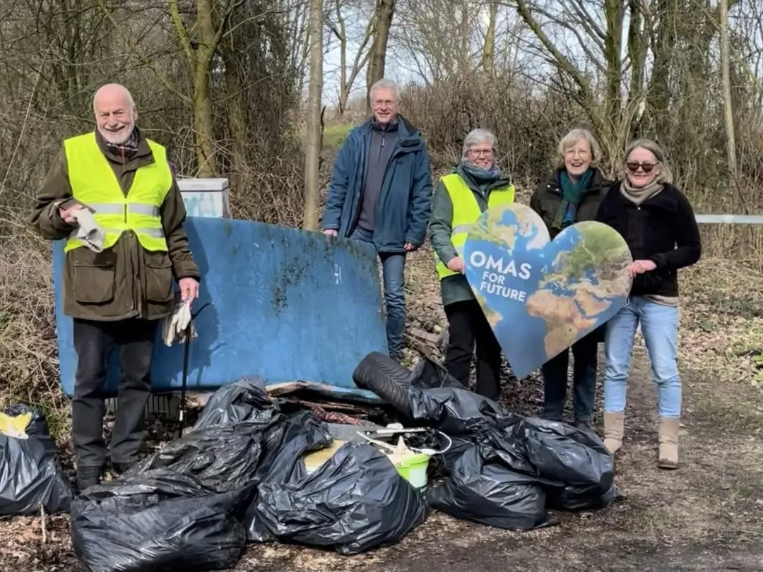 Fünf ältere Erwachsene stehen im Freien mit gesammeltem Müll in Tüten. Zwei tragen gelbe Sicherheitswesten, einer hält ein großes herzförmiges Schild mit der Aufschrift OMAS FÜR DIE ZUKUNFT. Im Hintergrund sind blattlose Bäume und Büsche zu sehen.