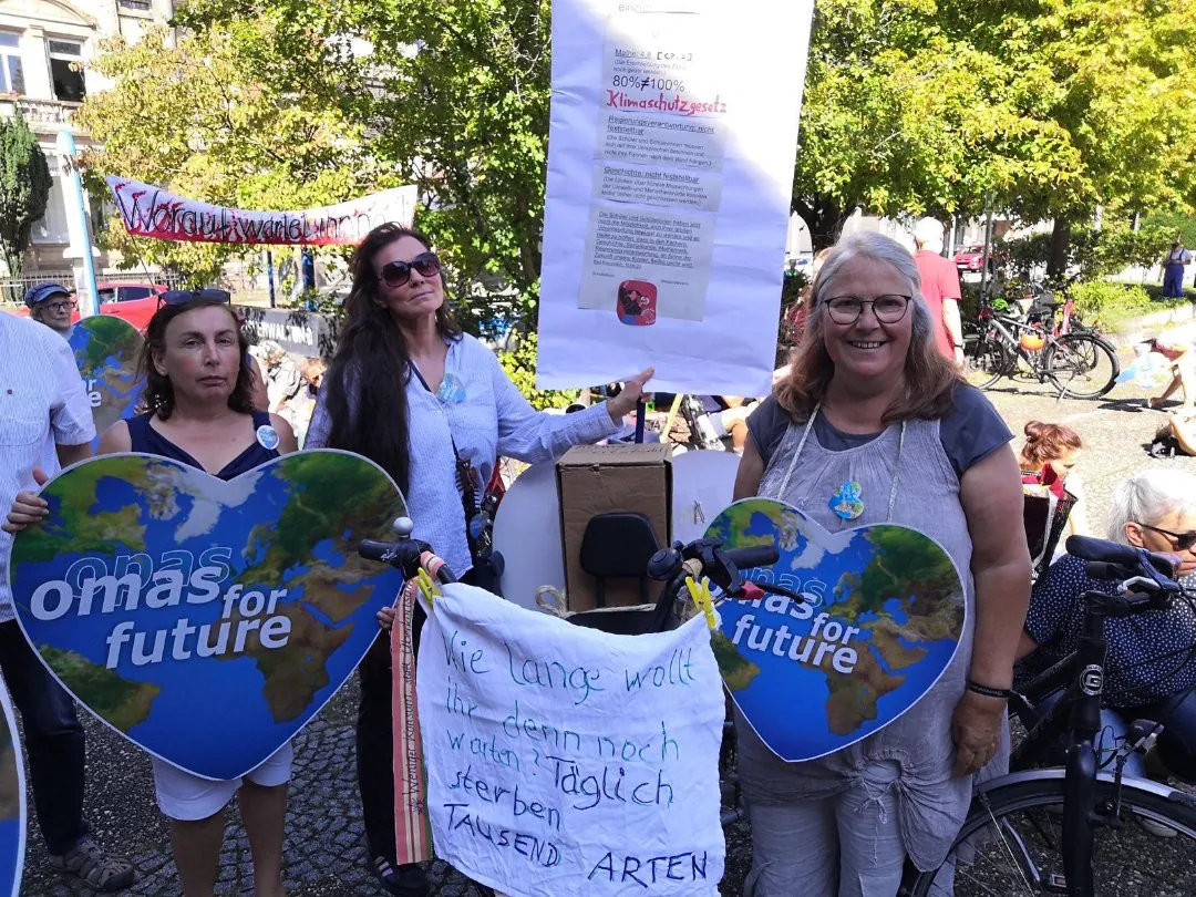 Drei Frauen halten bei einer Klimaprotestaktion herzförmige "Omas for Future"-Schilder und ein Transparent über das Artensterben. Über ihnen hängt ein Plakat mit Informationen. Andere Demonstranten und Fahrräder sind an einem sonnigen Tag im Hintergrund zu sehen.