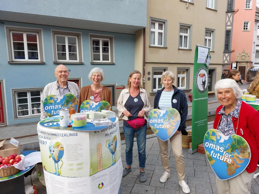 Fünf ältere Erwachsene stehen bei einer Klimaaktivismus-Veranstaltung im Freien zusammen und halten herzförmige Omas for Future-Schilder in die Höhe. Sie lächeln, und vor ihnen steht ein Tisch mit Informationsmaterial und Äpfeln. Im Hintergrund sind Gebäude zu sehen.