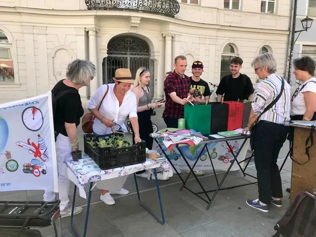 Eine Gruppe von Menschen versammelt sich um Tische, die auf einem Gehweg aufgestellt sind, und tauscht Pflanzen aus. Einige halten Tüten in der Hand, während andere getopfte Setzlinge arrangieren. Die Szene spielt sich vor einem weißen historischen Gebäude ab.