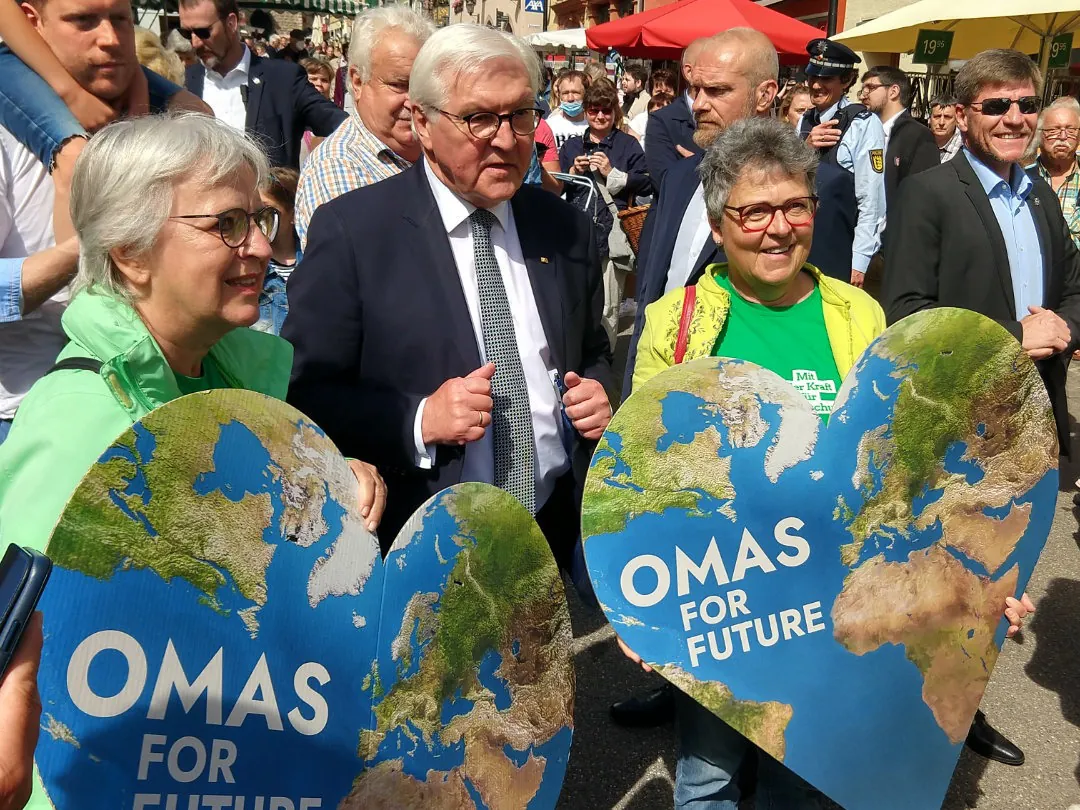 Zwei ältere Frauen halten herzförmige Schilder mit der Aufschrift OMAS FOR FUTURE (OMAS FÜR DIE ZUKUNFT) und einer Weltkugel auf einer überfüllten Veranstaltung im Freien. Ein Mann im Anzug und mit Brille steht zwischen ihnen; im Hintergrund sind Menschen und Polizei zu sehen.