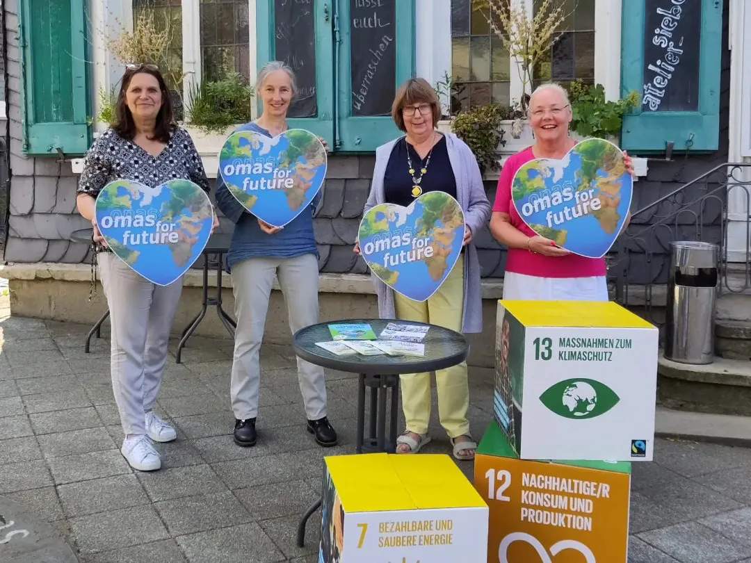 Vier Frauen stehen draußen und halten herzförmige Omas for Future-Schilder. Vor ihnen stehen Kästen mit den Zielen für nachhaltige Entwicklung in den Bereichen Klimaschutz, saubere Energie und verantwortungsvoller Konsum.