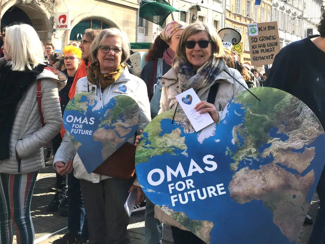 Zwei ältere Frauen halten bei einem Klimaprotest große herzförmige Schilder mit einer Weltkarte und dem Text OMAS FOR FUTURE. Im Hintergrund sind andere Menschen und Protestschilder zu sehen.