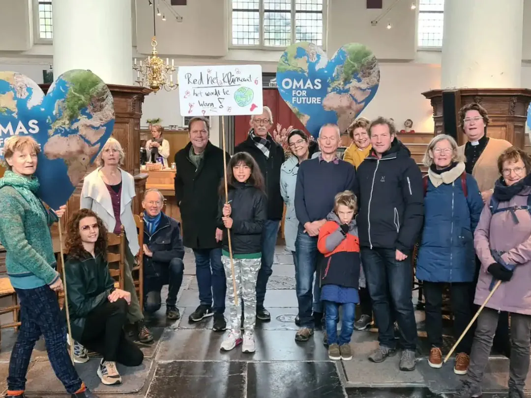 Eine Gruppe von Menschen unterschiedlichen Alters posiert in einem Innenraum mit Schildern und herzförmigen Kugeln. Auf einem Schild steht: "Real Net(Zero) would be... at whaat ke [Zukunft unklar]. Die Gruppe scheint Omas for Future zu unterstützen.