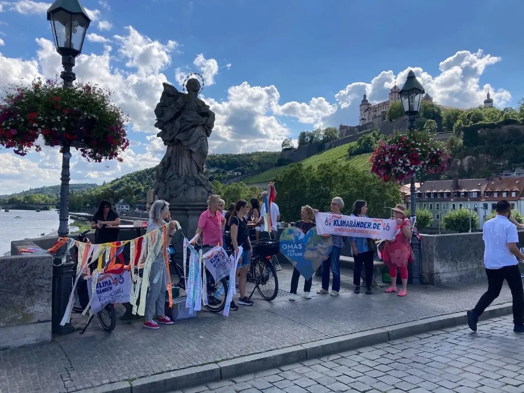 Eine Gruppe von Menschen steht an einem sonnigen Tag auf einer Brücke in der Nähe einer Statue und hält bunte Fahnen und Bänder in der Hand, während im Hintergrund Blumenkörbe und eine malerische Hügellandschaft zu sehen sind.