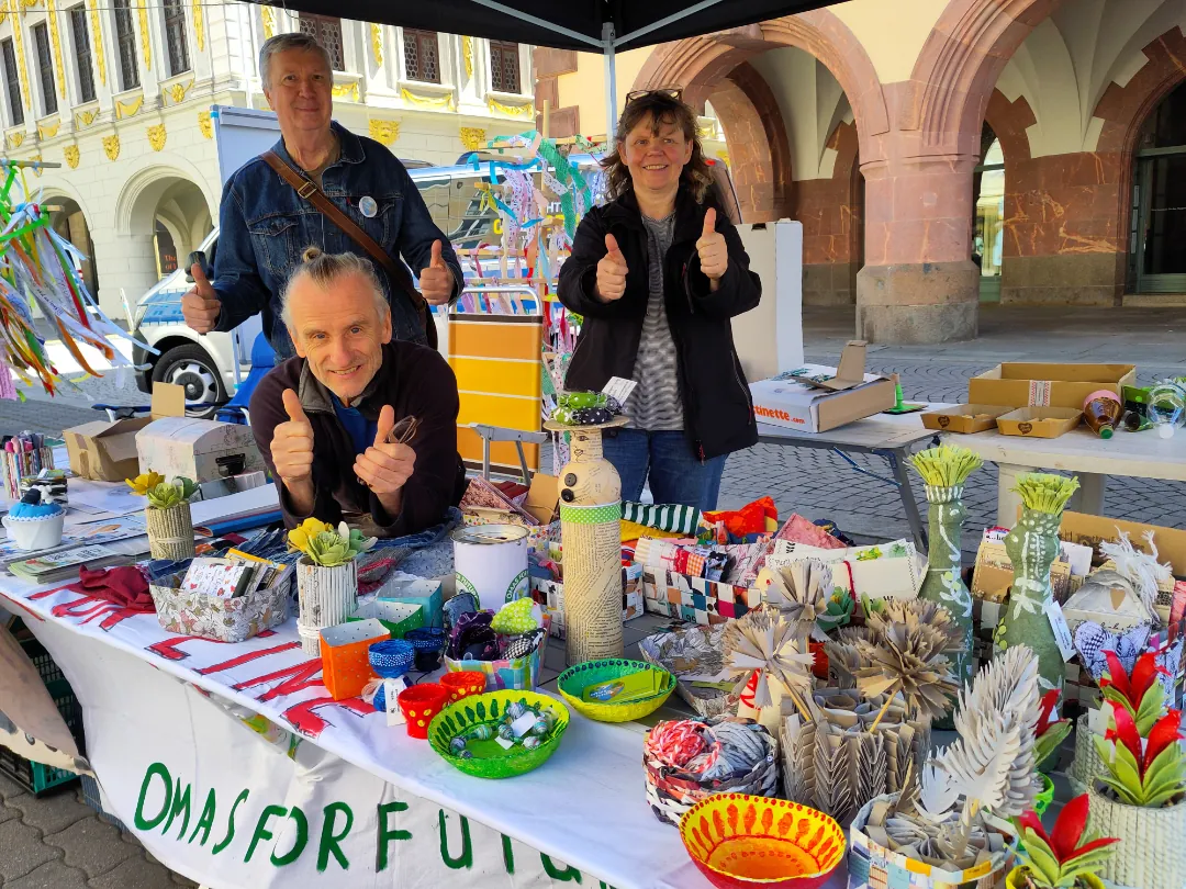 Drei lächelnde Erwachsene stehen hinter einem Marktstand mit buntem Kunsthandwerk und Origami. Zwei stehen, einer sitzt, alle zeigen den Daumen nach oben. Auf dem Schild des Standes steht OMAS FOR FUTURE. Hinter ihnen sind Gebäude und Dekorationen zu sehen.