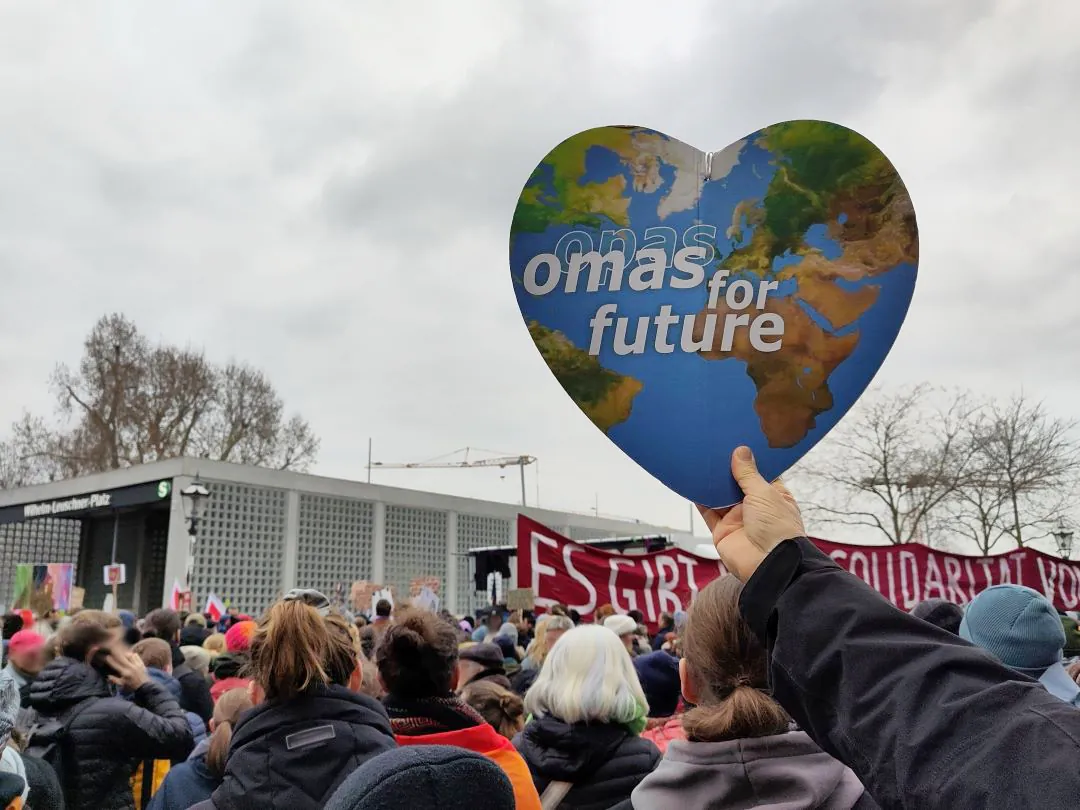 Eine Person hält bei einer großen Protestveranstaltung im Freien ein herzförmiges Schild mit der Aufschrift omas for future und einer Weltkarte in die Höhe. Unter bewölktem Himmel sind viele Menschen versammelt, einige halten Transparente.