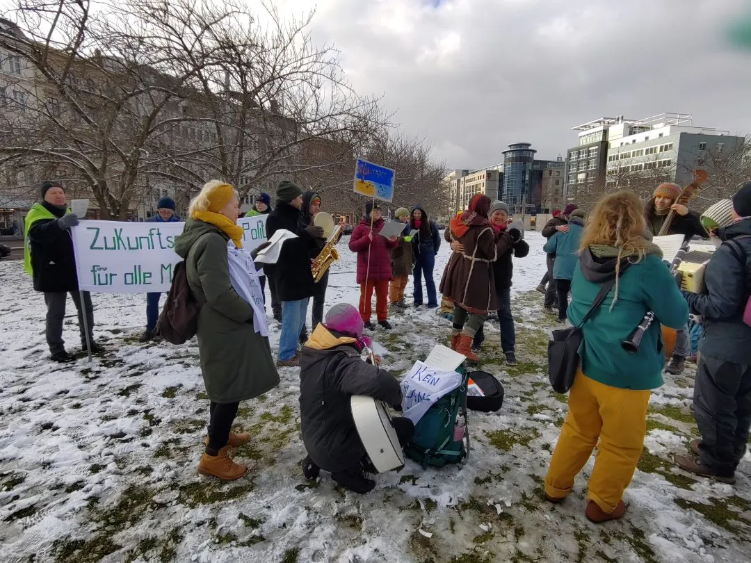 Eine Gruppe von Menschen steht mit Schildern und Musikinstrumenten auf einer verschneiten Wiese, um zu protestieren. Auf einem Transparent in deutscher Sprache steht "Zukunft...", und mehrere Personen sind in Winterkleidung gehüllt. Im Hintergrund sind Gebäude zu sehen.