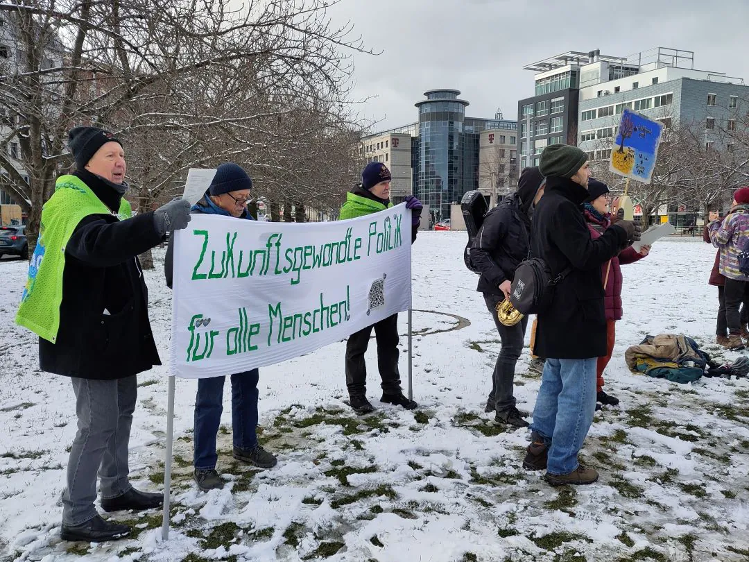 Eine Gruppe von Menschen steht auf verschneitem Gras und hält ein Banner mit der Aufschrift Zukunftsgewandte Politik für alle Menschen! in die Höhe. Einige tragen grüne Tücher, andere halten Schilder, im Hintergrund sind moderne Gebäude zu sehen.