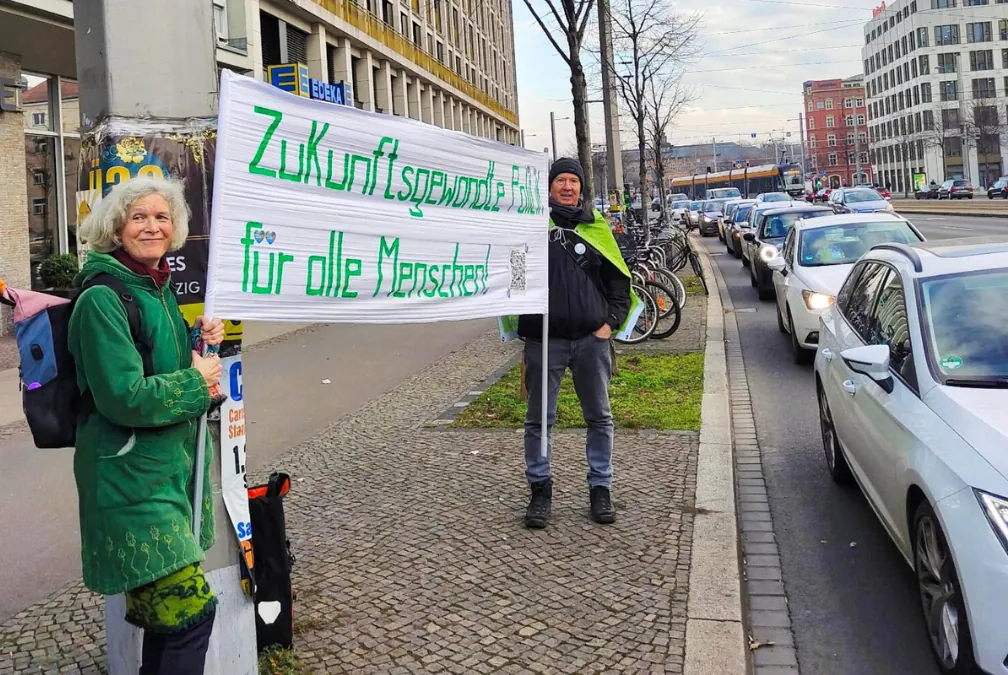 Zwei Personen stehen auf einem Bürgersteig an einer belebten Straße und halten ein großes Transparent mit der Aufschrift "Zukunftsgerechtigkeit für alle Menschen" in die Höhe. Im Hintergrund sind Autos und Gebäude zu sehen.