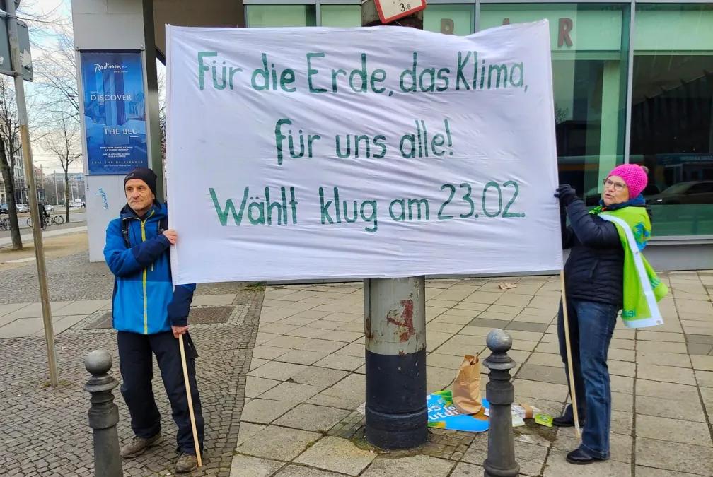 Zwei Personen stehen auf dem Bürgersteig einer Stadt und halten ein großes Transparent mit der Aufschrift "Für die Erde, das Klima, für uns alle! Wählt klug am 23.02." (Für die Erde, das Klima, für uns alle! Wählt klug am 23.02.).