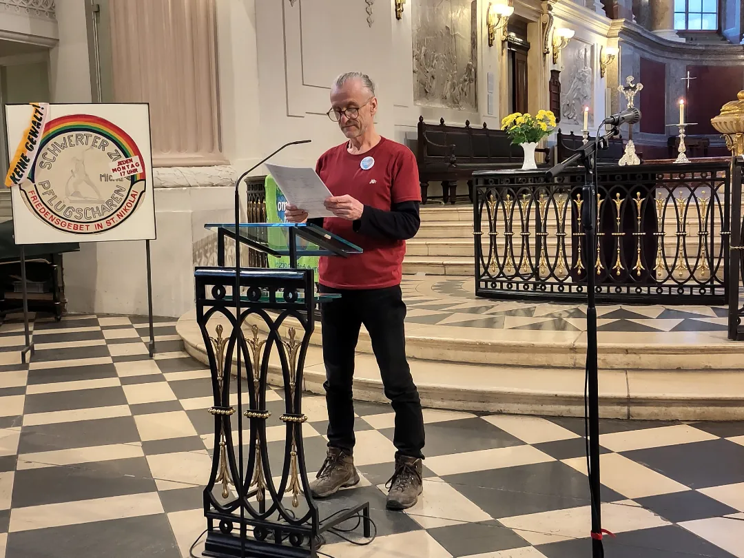 Ein Mann in einem roten Hemd steht an einem schwarzen Podium und liest in einer Kirche mit kariertem Boden von einem Blatt Papier ab. Hinter ihm steht ein buntes Schild und ein Altar mit Kerzen und gelben Blumen. In der Nähe ist ein Mikrofon aufgestellt.