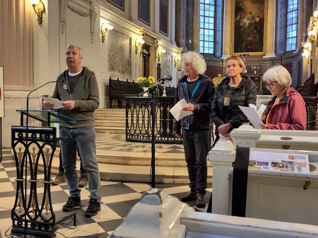 Vier Erwachsene stehen während eines Friedensgebets vor einer Kirche; ein Mann spricht an einem Podium, während die anderen Papiere halten. Der kunstvolle Innenraum ist mit Säulen, Kunstwerken und Blumen in der Nähe des Altars geschmückt.