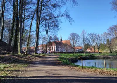 Ein unbefestigter Weg schlängelt sich durch einen Park mit hohen, blattlosen Bäumen am Rande eines Teiches. Im Hintergrund steht ein großes, hellrosa historisches Gebäude mit einem roten Dach und einem Glockenturm unter einem klaren blauen Himmel.