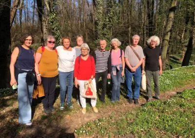 Eine Gruppe von neun Erwachsenen steht zusammen auf einem sonnenbeschienenen Waldweg, umgeben von Bäumen und grünem Laub. Sie sind lächelnd und leger gekleidet und genießen den Tag im Freien.