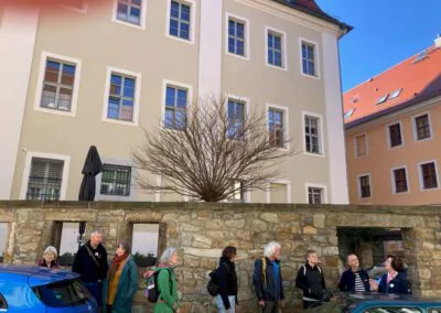 Eine Gruppe von Menschen steht und unterhält sich auf einer sonnigen Straße neben einer Steinmauer, mit einem blattlosen Baum und einem beigen Gebäude mit vielen Fenstern im Hintergrund. Einige von ihnen tragen Rucksäcke und Mäntel.