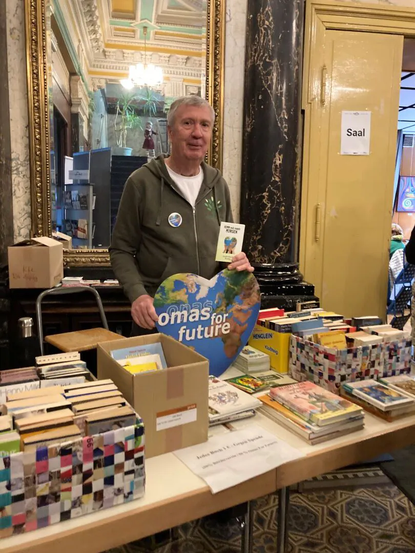 Ein älterer Mann steht bei einer Indoor-Veranstaltung hinter einem Tisch mit Büchern und einem herzförmigen Omas for Future-Schild. Im Hintergrund ist eine verschnörkelte Architektur mit Marmorsäulen und einer gelben Tür mit der Aufschrift Saal zu sehen.