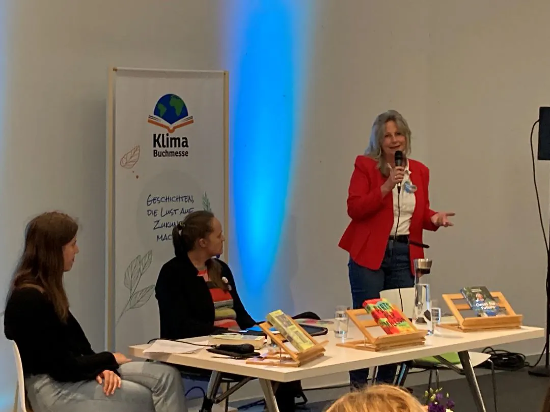 Drei Frauen nehmen an einer Podiumsdiskussion bei einer Indoor-Veranstaltung teil. Eine Frau steht und spricht in ein Mikrofon, während die beiden anderen an einem Tisch mit Büchern, Mikrofonen und einem Banner der Klima Buchmesse im Hintergrund sitzen.