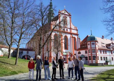 Eine Gruppe von Menschen steht und unterhält sich auf einem kopfsteingepflasterten Weg in der Nähe einer großen, verschnörkelten Kirche mit weißen und roten Wänden, hohen Fenstern und einem Turm, umgeben von blattlosen Bäumen unter einem klaren blauen Himmel.