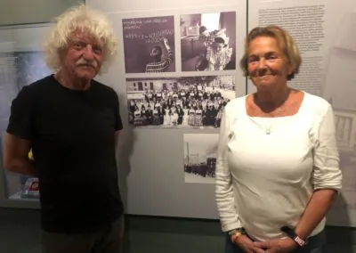 Zwei ältere Erwachsene, ein Mann und eine Frau, stehen vor einer Museumsausstellung mit historischen Schwarz-Weiß-Fotografien und informativem Text. Beide schauen in die Kamera und lächeln sanft.