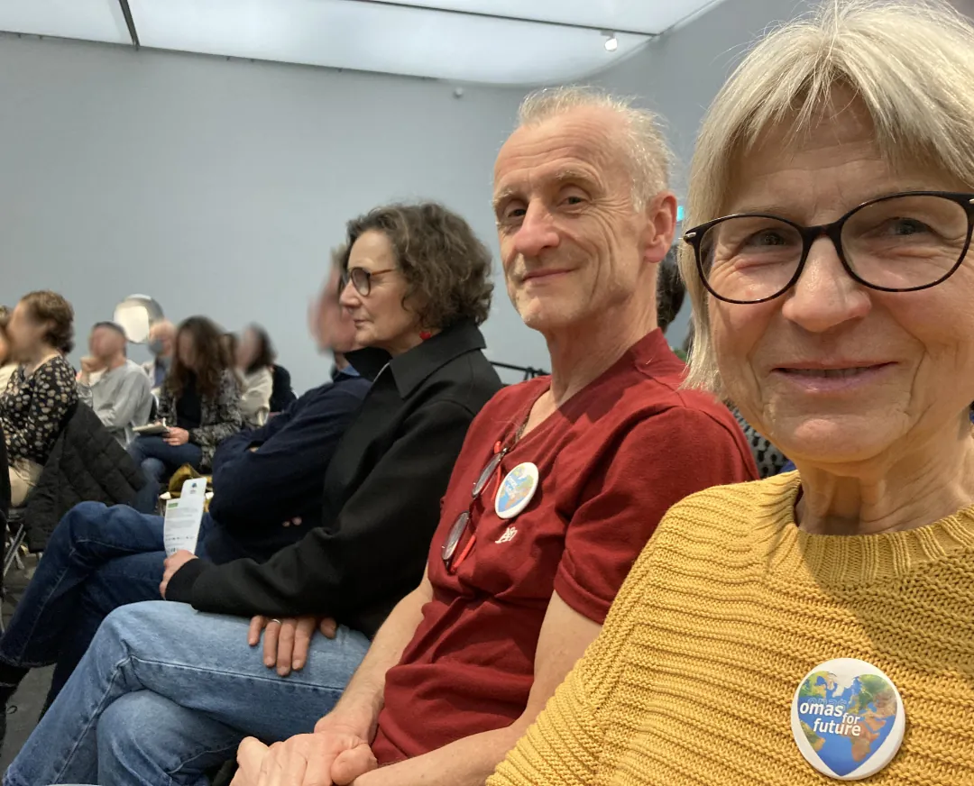 Eine Gruppe älterer Menschen sitzt lächelnd in einem gut beleuchteten Raum zusammen. Die Frau vorne trägt eine Brille und einen gelben Pullover mit einem runden Abzeichen, auf dem "omas" für "Zukunft" steht. Andere sitzen im Hintergrund, einige Gesichter sind verschwommen.