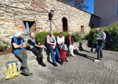 Eine Gruppe von sieben älteren Menschen sitzt und steht an einer sonnenbeschienenen Steinmauer, plaudert und entspannt sich auf einem Kopfsteinpflasterweg; einige haben Rucksäcke dabei und auch ein Hund ist dabei.