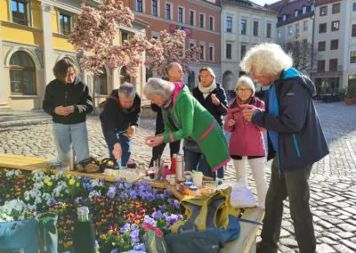 Eine Gruppe von Menschen versammelt sich an einem sonnigen Tag um ein Blumenbeet auf einer Kopfsteinpflasterstraße und teilt Essen und Getränke. In der Nähe blühen bunte Blumen, und im Hintergrund sind historische Gebäude mit blühenden Bäumen zu sehen.