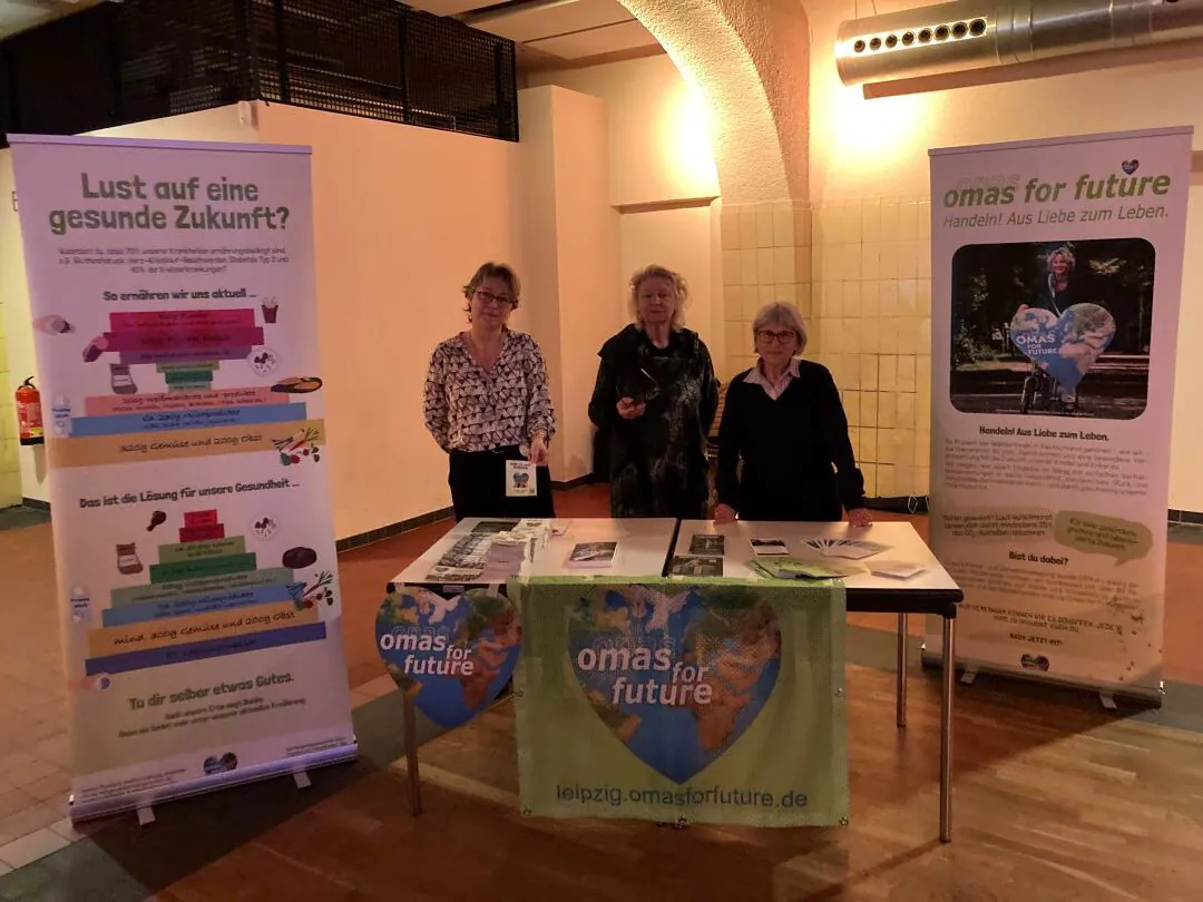 Drei Frauen stehen hinter einem Tisch mit Flugblättern und einem Transparent mit der Aufschrift "omas for future". Zwei große Plakate werben für Gesundheits- und Umweltaktionen. Die Szene scheint bei einer Veranstaltung in einem Innenraum mit warmer Beleuchtung zu spielen.