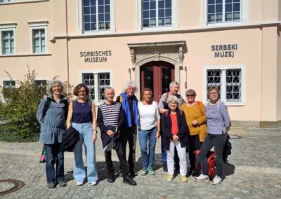 Eine Gruppe von neun Personen steht lächelnd vor dem Eingang des Sorbischen Museums, während das Sonnenlicht auf den kopfsteingepflasterten Weg und das helle Gebäude hinter ihnen scheint.