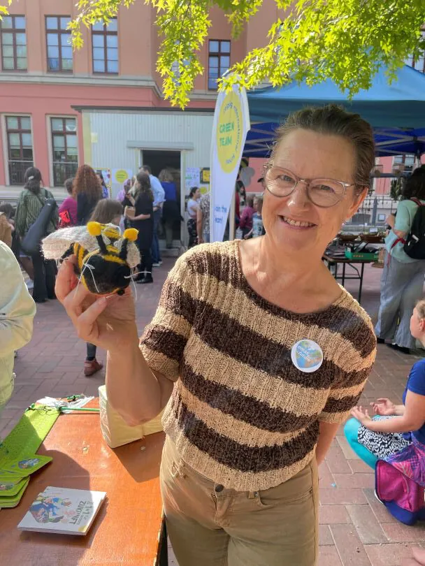 Eine lächelnde Frau mit Brille und gestreiftem Hemd hält eine kleine handgefertigte Bienenarbeit bei einer Veranstaltung im Freien hoch, mit Menschen und Ständen im Hintergrund. Das Sonnenlicht fällt durch die grünen Blätter der Bäume im Hintergrund.