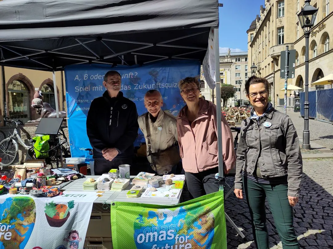Vier Personen stehen an einem Stand im Freien mit Informationsmaterial und einem Transparent in deutscher Sprache über ein Quiz zum Thema Zukunft. Der Stand zeigt grüne Tücher mit Omas für Zukunft und Bilder von der Erde. In der Nähe sind Gebäude und Fahrräder zu sehen.