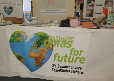 Ein Tisch mit einem Banner, auf dem "Omas for Future - Die Zukunft unserer Enkelkinder sichern" steht und das eine herzförmige Erdgrafik zeigt. Auf dem Tisch liegen gestrickte Artikel, Broschüren und Informationsmaterial.