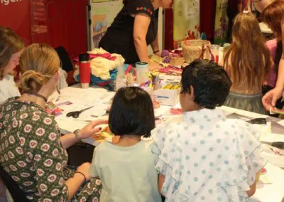 Kinder und Erwachsene versammeln sich bei einer Indoor-Veranstaltung um einen Tisch mit bunten Bastelmaterialien und beschäftigen sich mit Kunst und Handwerk. An den Wänden im Hintergrund hängen Plakate.