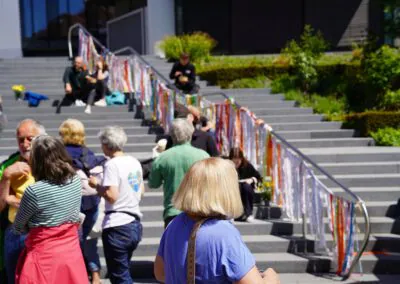 Eine Gruppe von Menschen versammelt sich an einem sonnigen Tag im Freien auf einer Treppe, die mit bunten Bändern geschmückt ist, die entlang des Geländers hängen, mit Grünflächen und Gebäuden im Hintergrund.