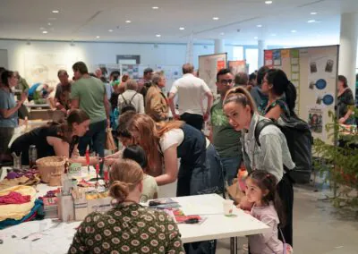 Eine geschäftige Indoor-Veranstaltung mit vielen Menschen, die um Tische mit Bastelarbeiten, Papieren und Materialien herumstehen und sich austauschen. Im Hintergrund sind bunte Auslagen und Informationsplakate aufgestellt.