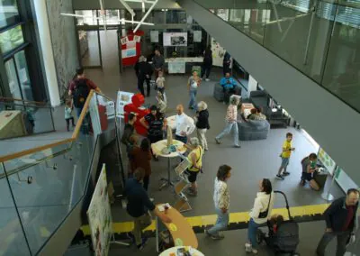 Ein belebter überdachter öffentlicher Raum, in dem Erwachsene und Kinder miteinander kommunizieren, Auslagen betrachten und umhergehen. Einige Menschen stehen an Informationsständen, andere sitzen oder unterhalten sich in Gruppen. Auf der linken Seite sind Treppen und ein Glasgeländer zu sehen.
