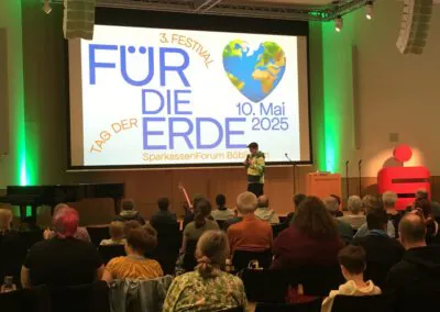 Ein Redner wendet sich an das Publikum in einer modernen Halle, hinter ihm ein großer Bildschirm mit der Aufschrift FÜR DIE ERDE, 10. Mai 2025, SparkassenForum Böblingen und eine Weltkugelgrafik. Die Menschen sitzen mit Blick auf die Bühne.