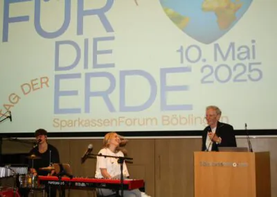 Ein Mann spricht an einem Podium mit der Aufschrift SparkassenForum, während zwei Musiker, einer am Schlagzeug und einer am Keyboard, auf der Bühne lächeln. Auf einer großen Leinwand hinter ihnen steht Tag der Erde 10. Mai 2025.