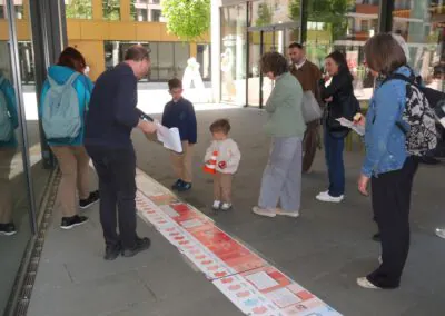 Eine Gruppe von Erwachsenen und Kindern steht draußen und betrachtet eine lange Zeitleiste oder ein Display, das auf dem Boden ausgelegt ist. Ein Mann deutet mit einer Geste darauf, während ein Kind in der Mitte sie genau betrachtet. Im Hintergrund sind Gebäude und Bäume zu sehen.