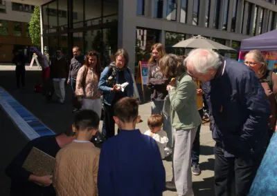 Eine Gruppe von Erwachsenen und Kindern versammelt sich an einem sonnigen Tag im Freien und schaut auf etwas auf dem Boden herab. Sie sind umgeben von modernen Gebäuden und bunten Ständen.