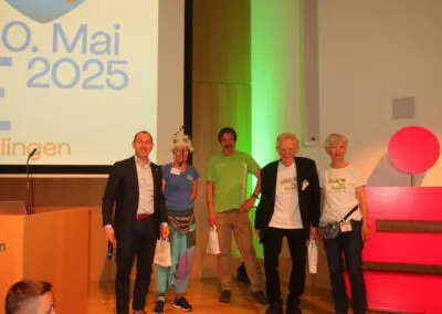 Fünf Personen stehen lächelnd auf einer Bühne bei einer Veranstaltung, einige halten Geschenktüten. Auf einem Präsentationsbildschirm hinter ihnen wird der 10. Mai 2025. Eine Person trägt ein buntes Kostüm mit einer weißen Perücke und einem Hut.