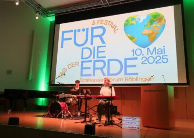 Zwei Musiker spielen auf der Bühne mit Schlagzeug und Keyboard vor einem großen Bildschirm, auf dem 3. Festival Für Die Erde, 10. Mai 2025, Tag der Erde, Sparkassen Forum Böblingen und eine Erdgrafik.