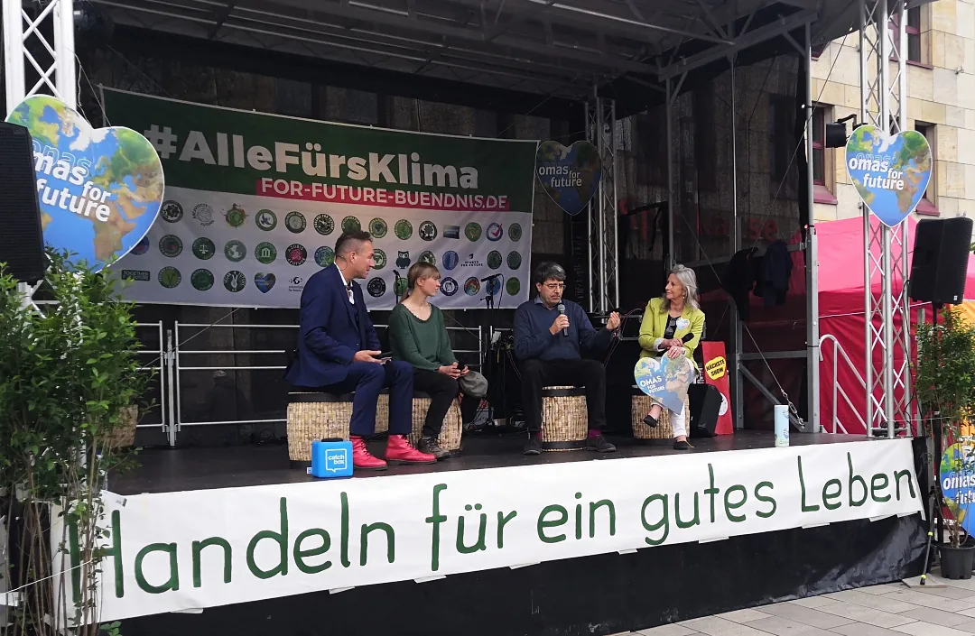 Vier Personen sitzen auf einer Bühne mit Mikrofonen während einer Klimaveranstaltung. Auf einem Banner hinter ihnen steht #AlleFürsKlima. Auf einem anderen Transparent davor steht Handeln für ein gutes Leben.