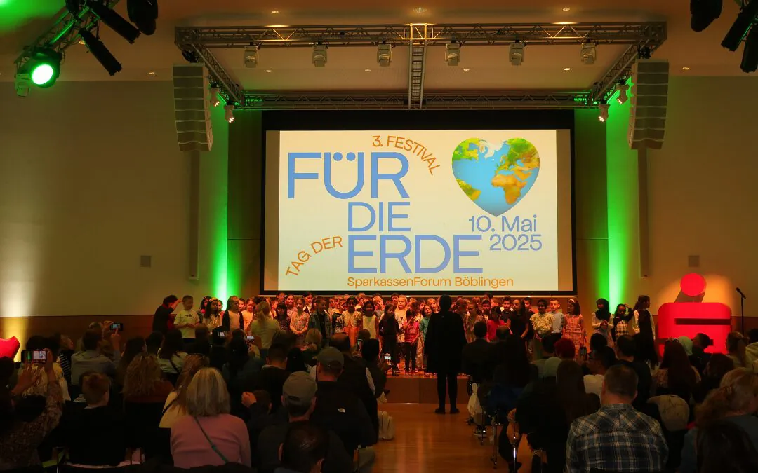 Ein großes Publikum beobachtet Kinder, die auf der Bühne vor einer Leinwand mit der Aufschrift Für die Erde, einer herzförmigen Erde und dem Datum 10. Mai 2025 im SparkassenForum Böblingen, mit grüner Beleuchtung an den Wänden.